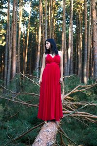 Full length of woman wearing red dress standing on fallen tree in forest