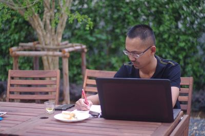 Man writing in book while sitting at table against plants