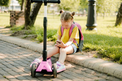 A smiling little schoolgirl girl is sitting on the curb with a phone in her hands during a break 