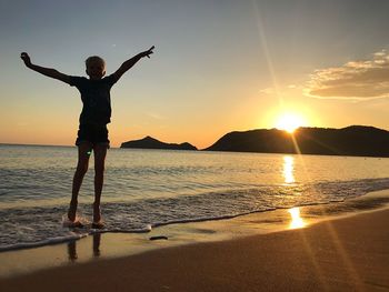 Boy with arms outstretched standing at beach during sunset