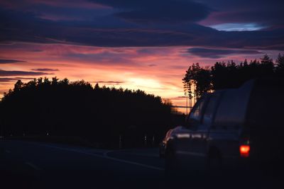 Silhouette trees by road against sky at sunset