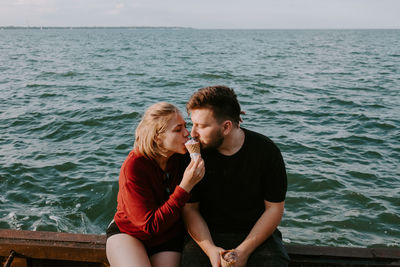 Happy young romantic couple in casual wear eating ice cream cone together while sitting on pier near sea and enjoying summer holidays together