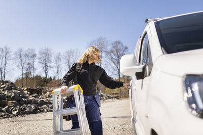 Female electrician with ladder opening van's door standing on sunny day
