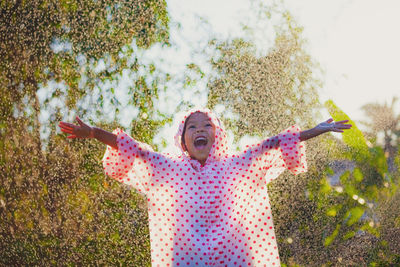 Happy girl wearing raincoat with arms outstretched standing in park during rainfall