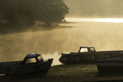 Boats moored on shore beside a backlit mist covered lake