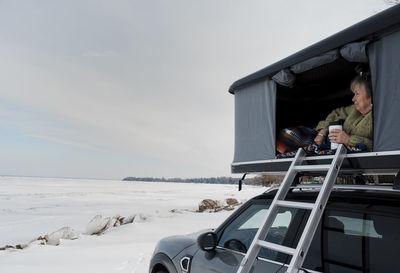 Woman looking away while relaxing in roof tent on car against cloudy sky