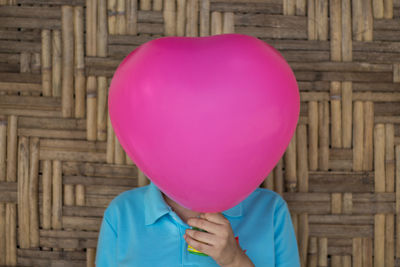 Woman covering face with pink balloon against wooden wall