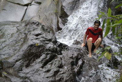 Low angle view of young man sitting on rock in waterfall