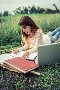 Young woman writing in book by laptop on field