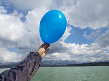 Cropped hand holding balloon over lake against sky