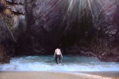 Rear view of shirtless man enjoying on shore against rock formations