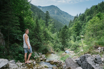 Portrait of smiling young man standing on rock by stream in forest