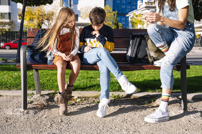 Low section of mother with kids using smart phone while sitting on bench in public park during sunny day