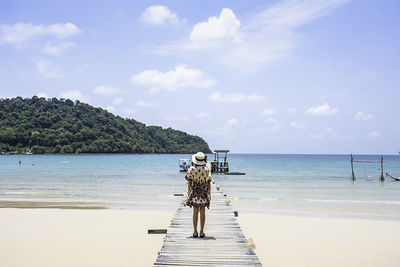 Rear view of woman wearing hat standing on pier at beach against sky