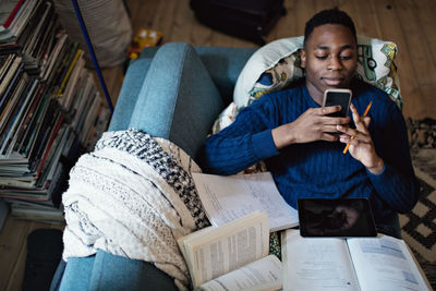 High angle view of teenage boy using mobile phone while lying on sofa with books at home