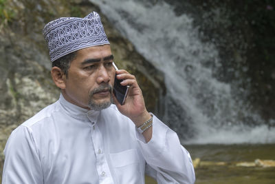 Thoughtful man talking on phone while sitting against waterfall