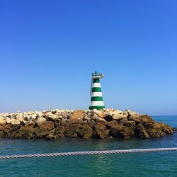 Lighthouse by sea against clear blue sky