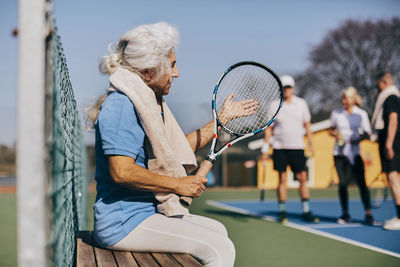 Senior woman holding tennis racket while sitting on bench at tennis court