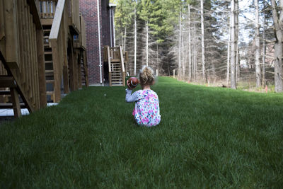 Rear view of girl holding american football while sitting on grassy field in yard