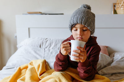 Child wearing grey warm hat sits on the bed under warm blanket and drinks hot tea