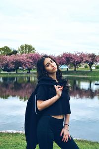 Beautiful young woman using phone while standing by lake against sky