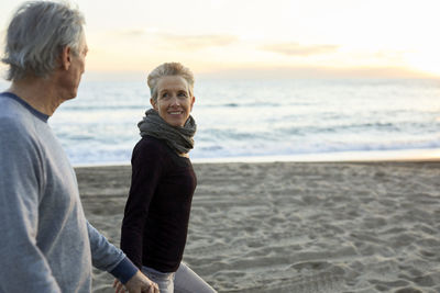 Side view of senior couple holding hands while walking at beach during sunset