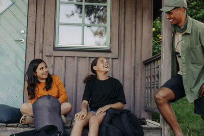 Smiling girls sitting while talking to male counselor near cabin at summer camp