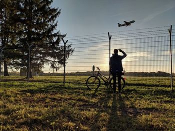 Silhouette man with bicycle waving hand at airplane flying in sky