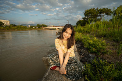 Portrait of smiling young woman sitting by plants against sky