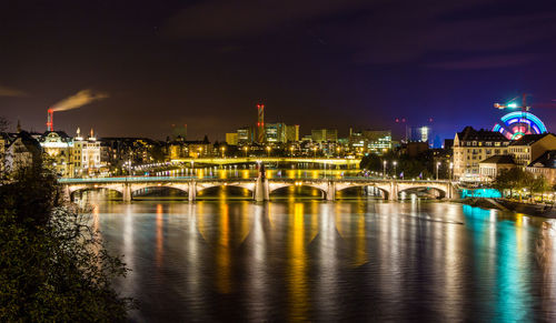 Illuminated bridge over river by buildings against sky at night