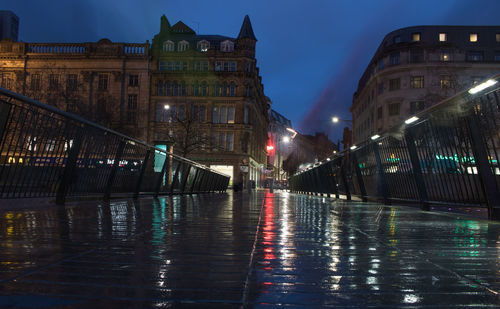 Wet illuminated street by buildings in city at night