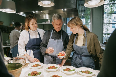 Chef instructing male and female students while plating during cooking class in kitchen