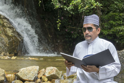 Man reading book while sitting against waterfall