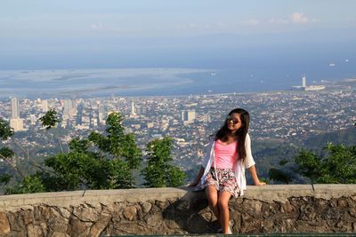 Smiling young woman sitting on stone wall in city against sky