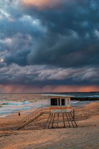 Scenic view of beach and life saver hut against stormy sky during sunset