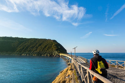 Man walking on bridge over sea towards island against sky