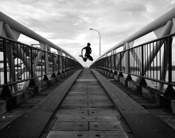 Full length rear view of man jumping on footbridge against sky