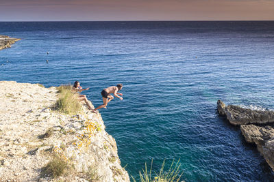 People jumping on rock by sea against sky