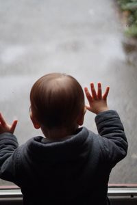 Rear view of curious boy looking through wet window during rainy season