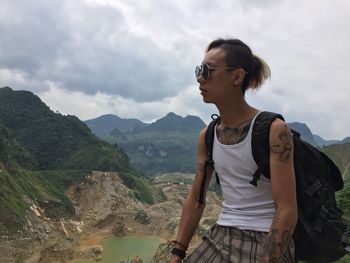 Young woman with backpack standing on mountain against cloudy sky