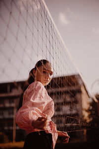 Side view portrait of young woman standing by volleyball net against blue sky