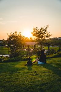 Rear view of people sitting on field against sky during sunset