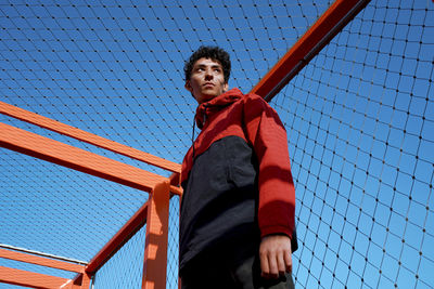 Thoughtful young man standing under fence against clear blue sky