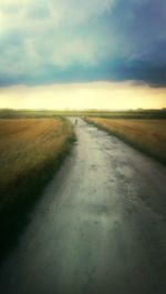 Road passing through field against cloudy sky