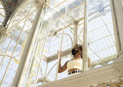 Trendy ethnic female with braids wearing protective mask standing in glass building lit by sunlight while looking through window