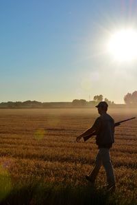 Full length of man holding rifle while walking on grass against sky during sunset