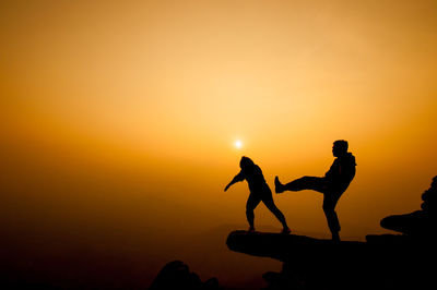 Silhouette man kicking woman from cliff against sky during sunset