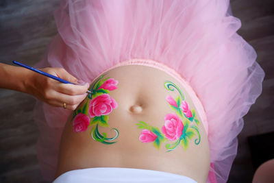 Close-up of a pregnant belly, on which the artist paints with a brush aqua makeup of pink flowers