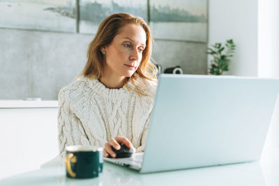 Young adult forty years beautiful blonde woman with long hair working on laptop sitting on kitchen 
