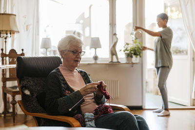 Elderly woman sitting on chair and knitting yarn while female nurse watering plants in background at home